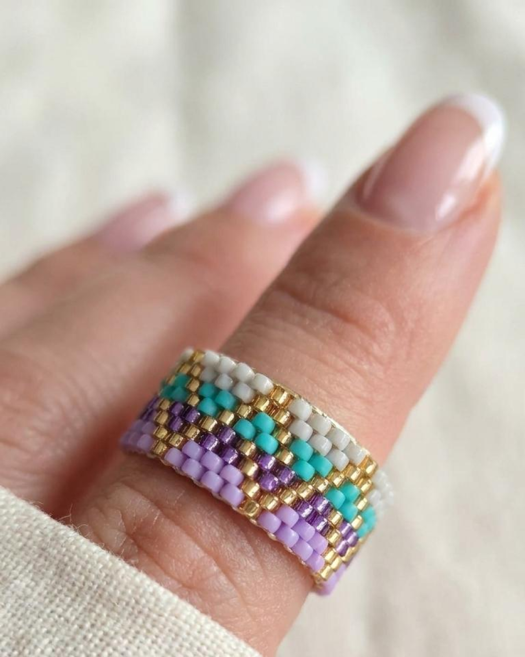 Hand wearing a woven Miyuki ring in lilac, aqua, white, and gold tones.