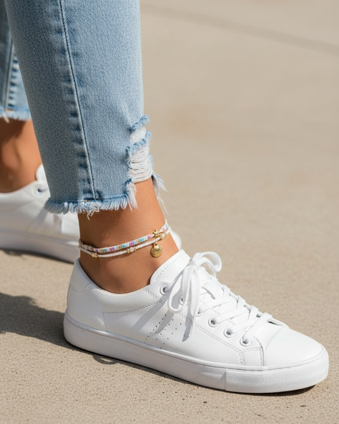 Double anklet with pastel beads and gold shell charm, worn with white sneakers and frayed jeans in sunlight.