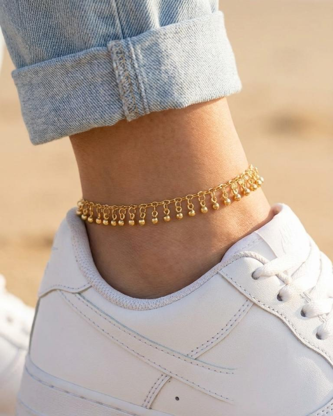 Gold anklet with dangling charms styled with white sneakers and light-wash jeans.