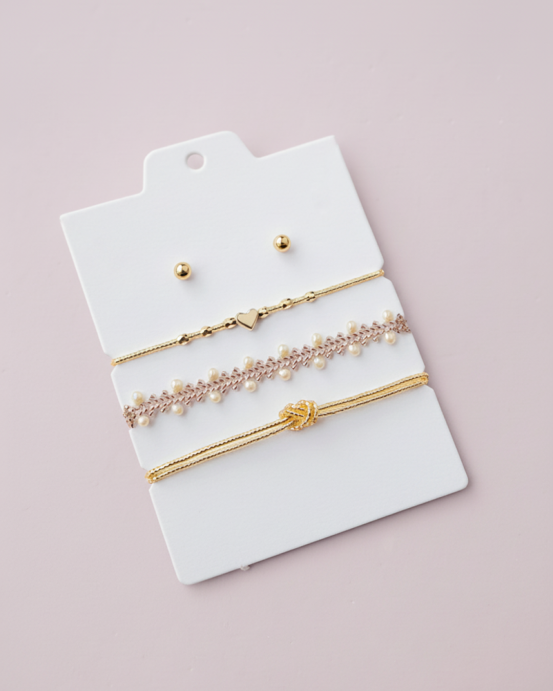 Bracelet trio (gold heart, lace with pearls, knot) and gold studs on a white display card, pink backdrop.