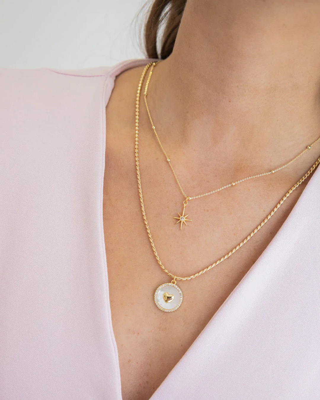 Layered set of two gold necklaces: fine chain with star pendant and twisted chain with heart medallion over a V-neck.