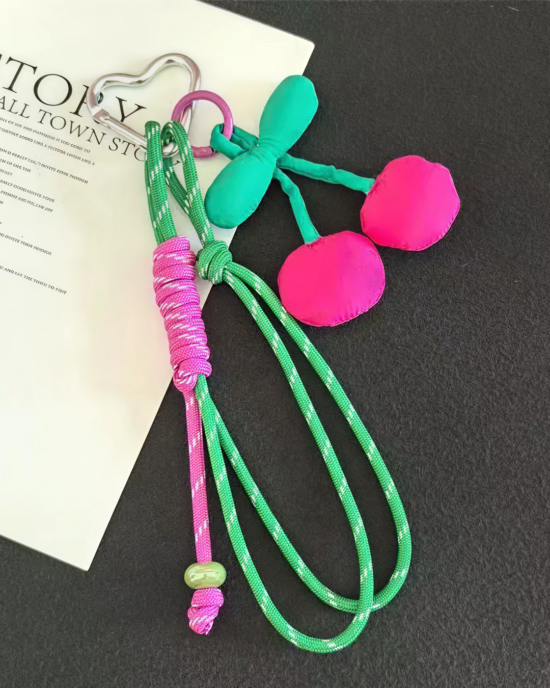 Cherry bag charm with fuchsia padded cherries, green cord, and silver heart carabiner on a dark background.