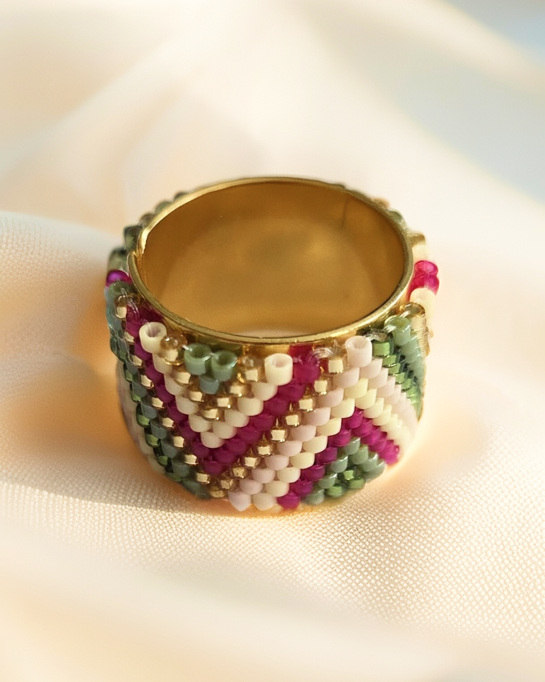Wide band ring handwoven with Miyuki beads in a green, fuchsia, and cream chevron pattern on a satin backdrop, macro view.