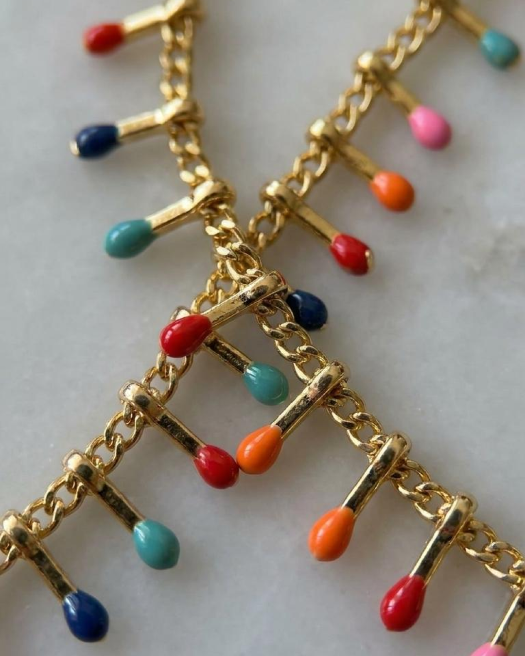 Macro detail of a gold anklet with enamel drop charms in red, blue, pink, orange, and turquoise.