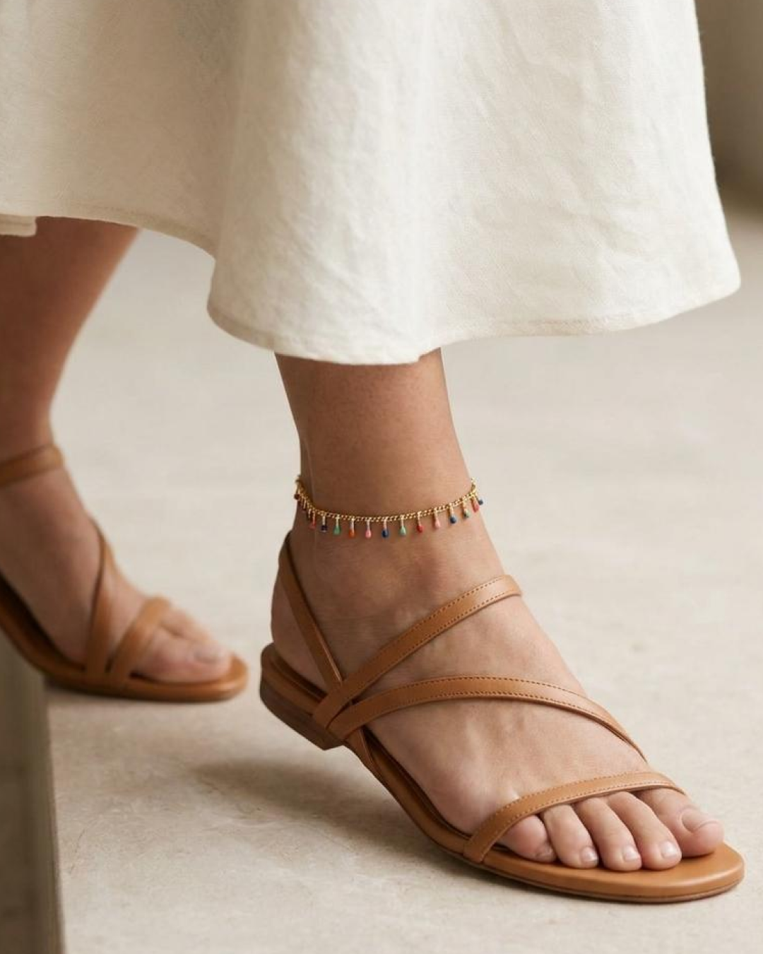Gold anklet with tiny multicolor drop charms worn on the ankle, featuring an adjustable chain.