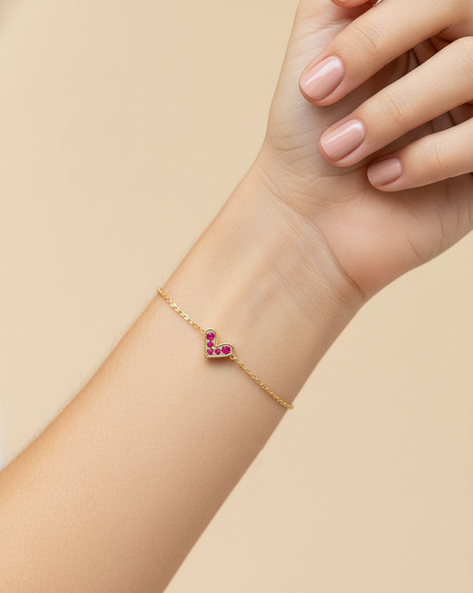 Gold-plated stainless steel bracelet with fuchsia crystal heart charm on wrist against beige background.