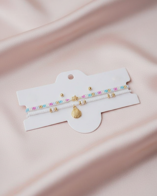 Double anklet set on white card: pastel strand with stars, white strand with spacers, and a gold shell charm.