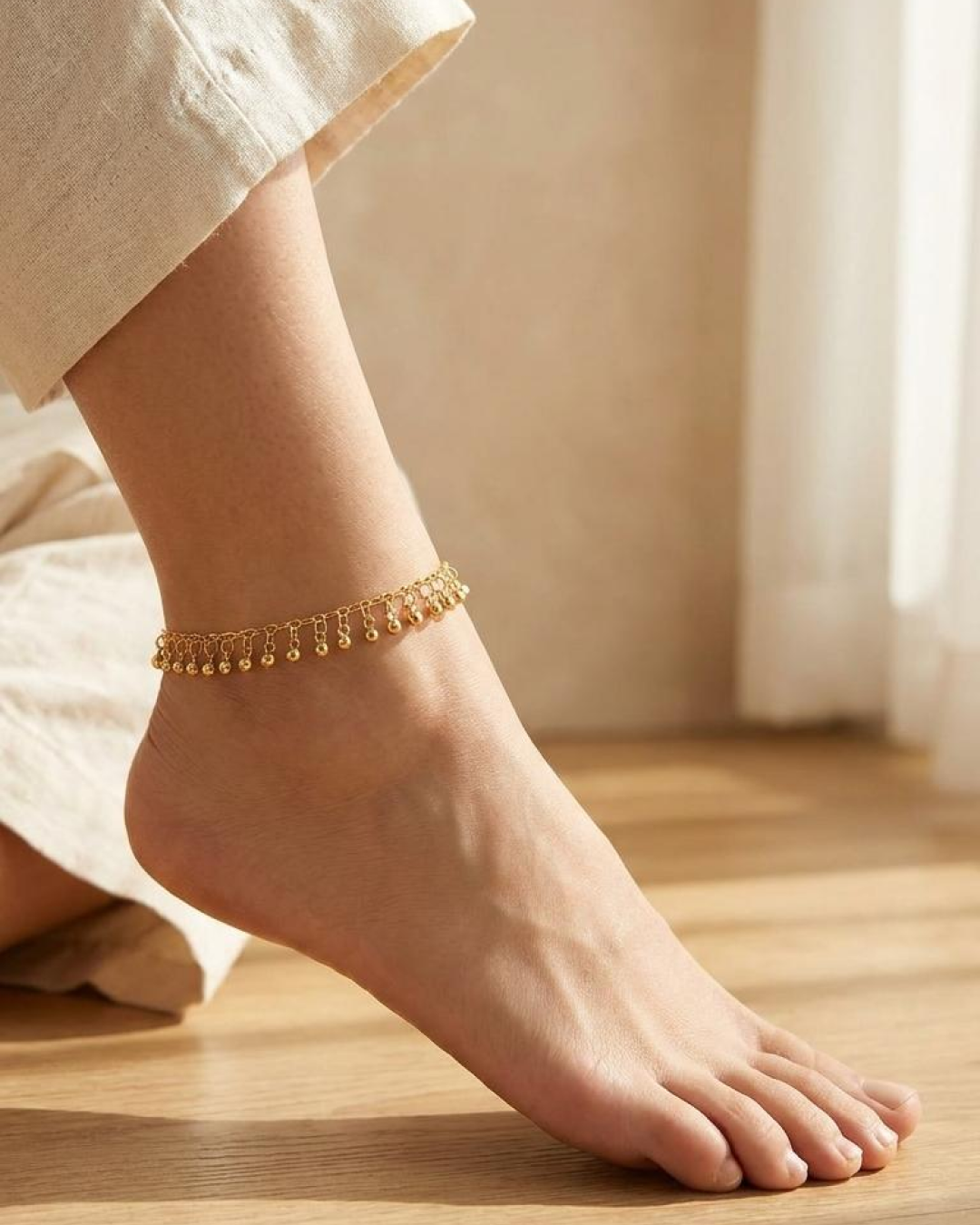 Delicate gold anklet worn on the ankle with light-colored trousers in soft indoor light.