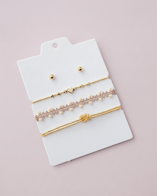 Bracelet trio (gold heart, lace with pearls, knot) and gold studs on a white display card, pink backdrop.