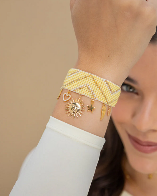 Wide yellow woven bracelet made with Czech seed beads, gold spacers and goldfilled charms (heart, sun, star and feather) with sliding-knot closure on a woman’s wrist.