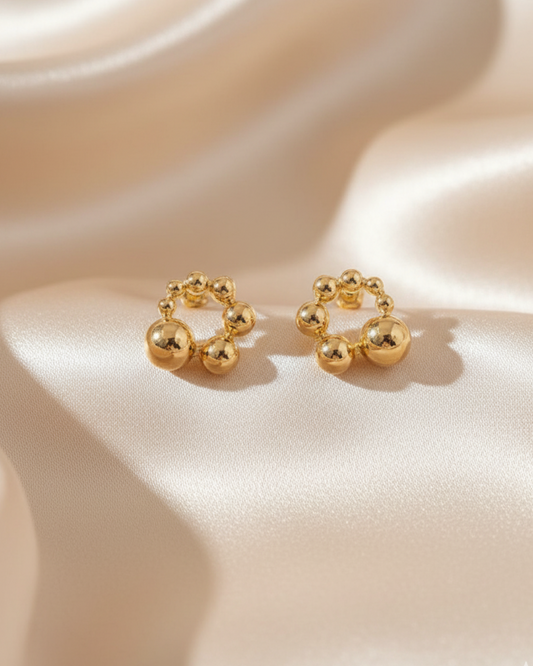 Pair of gold bubble stud earrings forming small hoops on a beige satin background with soft lighting.