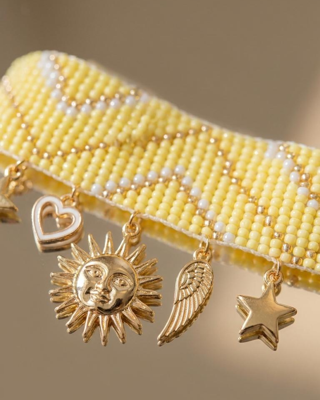 Close-up of a handwoven yellow beaded bracelet with gold-tone dangling charms, including a heart, sun, wing, and star.