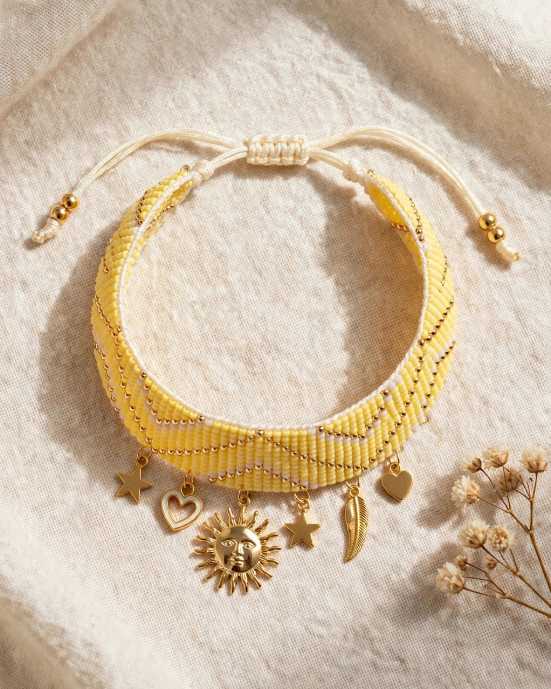 Handwoven yellow beaded bracelet with gold-tone sun, star, heart, and feather charms, featuring an adjustable sliding-knot closure, displayed on beige linen fabric.