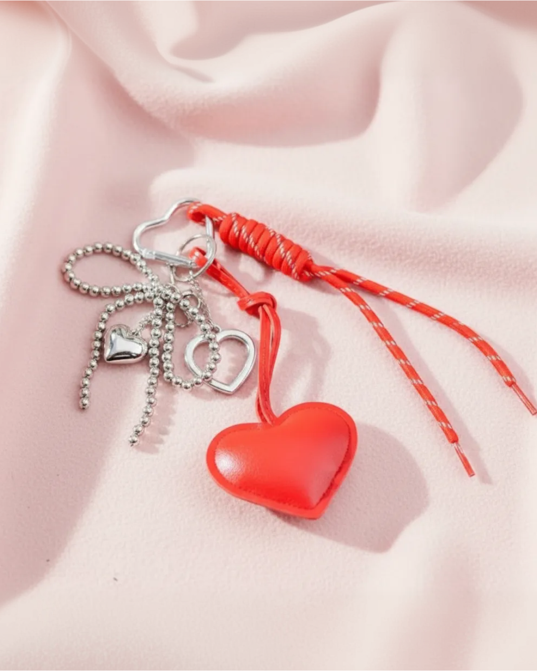 Red heart bag charm with silver mini hearts, ball chain and red cord, flat lay on soft pink fabric.