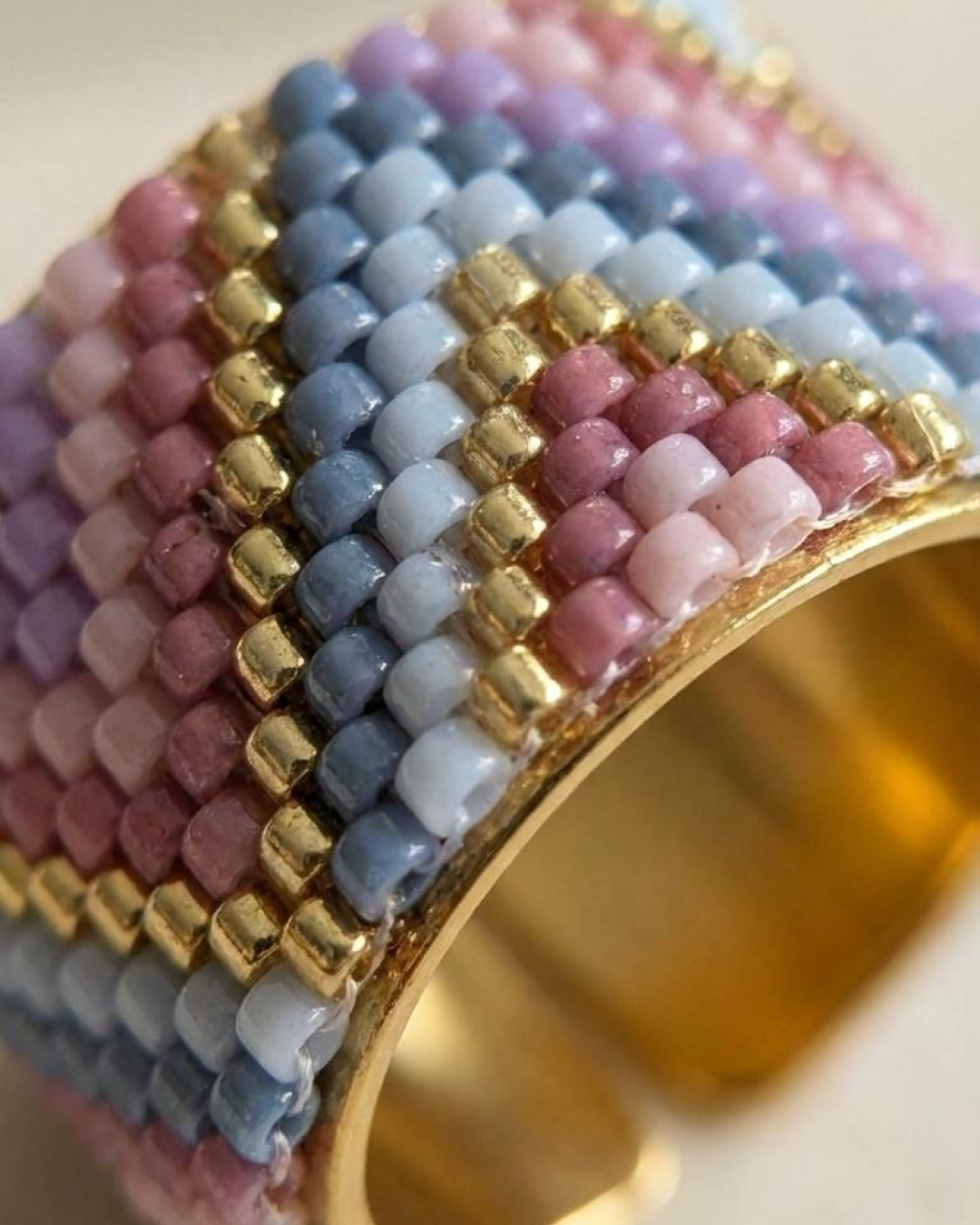 Close-up of a hand wearing a Miyuki-woven ring in lilac, pink, and blue on beige fabric.