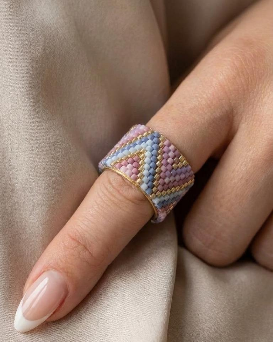 Close-up of a hand wearing a Miyuki-woven ring in lilac, pink, and blue on beige fabric.