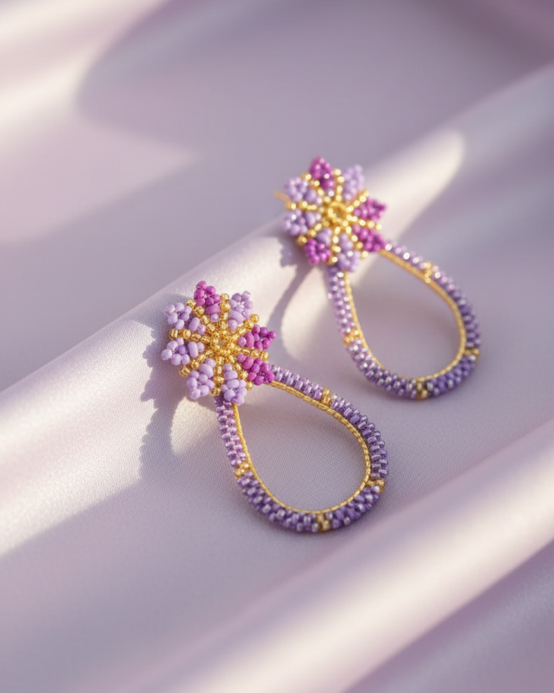 Pair of lilac flower and teardrop Miyuki earrings on lilac fabric, gold details.