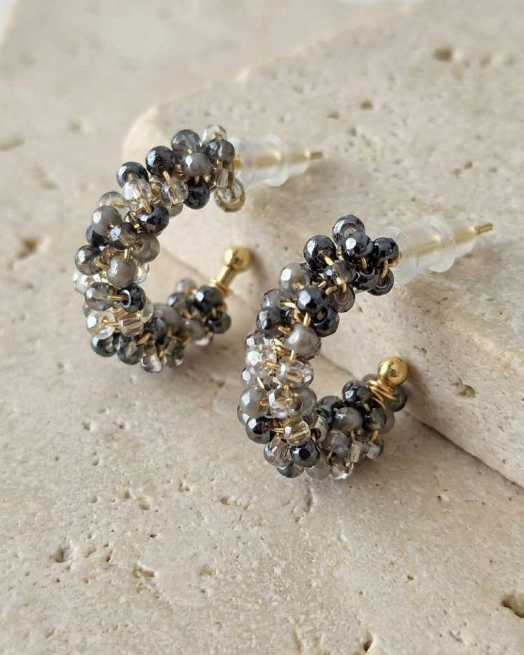 Pair of grey beaded mini hoop earrings with a gold-tone base displayed on a textured stone surface.