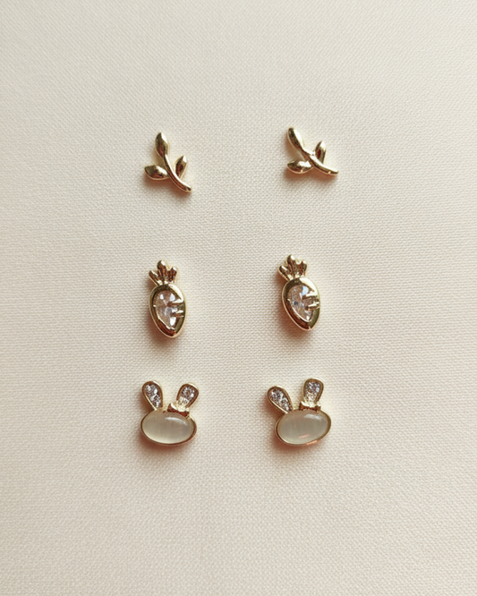 Mini gold-plated steel stud trio: leaves, carrot studs with cubic zirconia, and bunnies on a beige background.