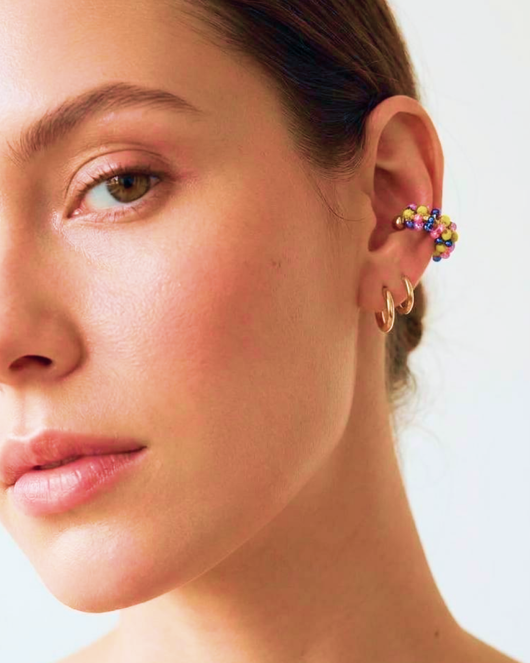 Portrait of a woman wearing a vibrant multicolor beaded ear cuff styled with small gold hoop earrings.