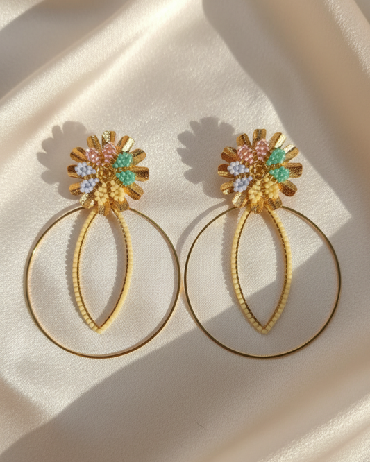 Pair of pastel Miyuki flower earrings with large golden hoops on beige satin fabric.