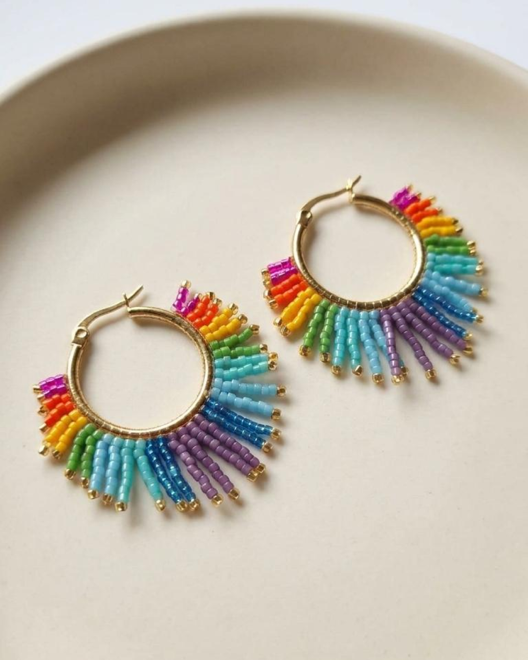 Pair of gold hoop earrings with rainbow-effect Miyuki fringe displayed on a white tray.