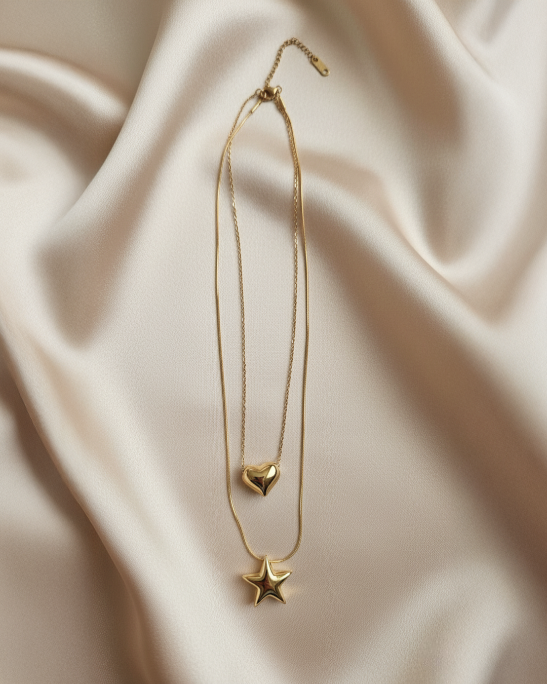 Two gold chains with heart and star pendants joined by a single clasp, flatlay on light satin.