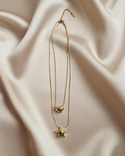 Two gold chains with heart and star pendants joined by a single clasp, flatlay on light satin.