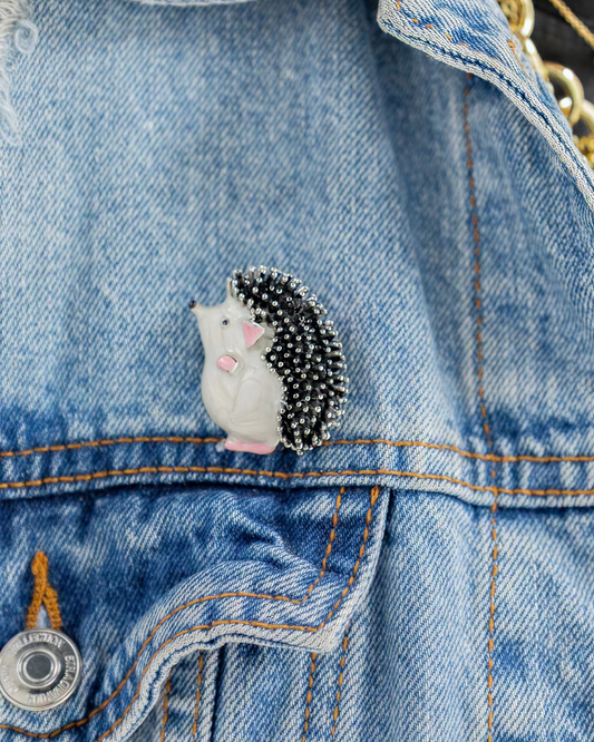 Hedgehog brooch pin attached to a blue denim jacket, featuring black and silver beaded spikes, a white body, and pink accents for a playful everyday look.