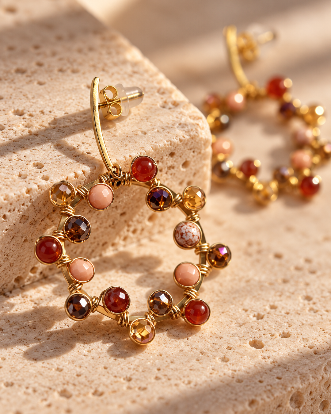 Close-up of a gold-plated circular drop earring decorated with red, amber, blush, and brown beads on a textured stone surface.