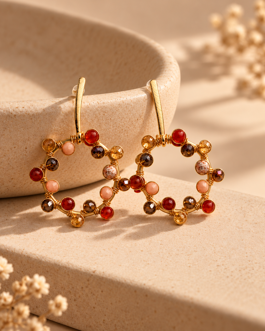 Close-up of a gold-plated circular drop earring decorated with red, amber, blush, and brown beads on a textured stone surface.