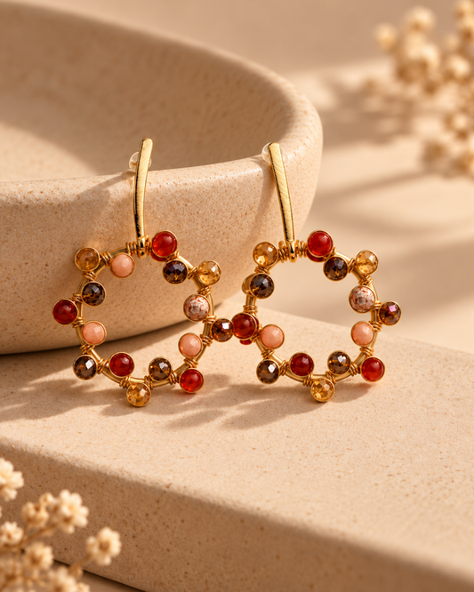 Close-up of a gold-plated circular drop earring decorated with red, amber, blush, and brown beads on a textured stone surface.