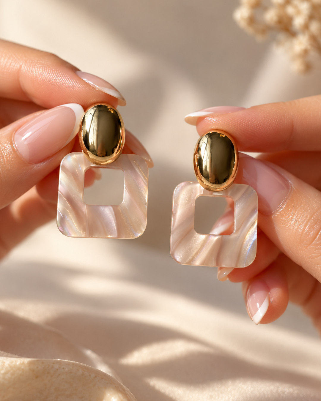 Close-up of Rosé Frame earrings held between fingertips, highlighting their glossy gold oval tops and translucent pink resin square pendants.