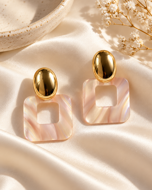 Pair of Rosé Frame statement earrings with polished gold oval studs and open square blush pink resin drops displayed on cream satin.